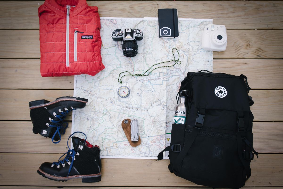 black hiking backpack near white Fujifilm instax mini camera near black leather boots, red half-zip jacket, gray pocket watch on white map.