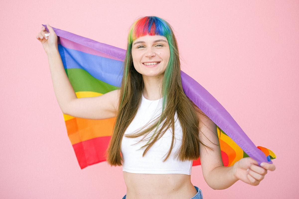 Young woman with rainbow fringe holds the pride flag. She wears a white crop top and is smiling.