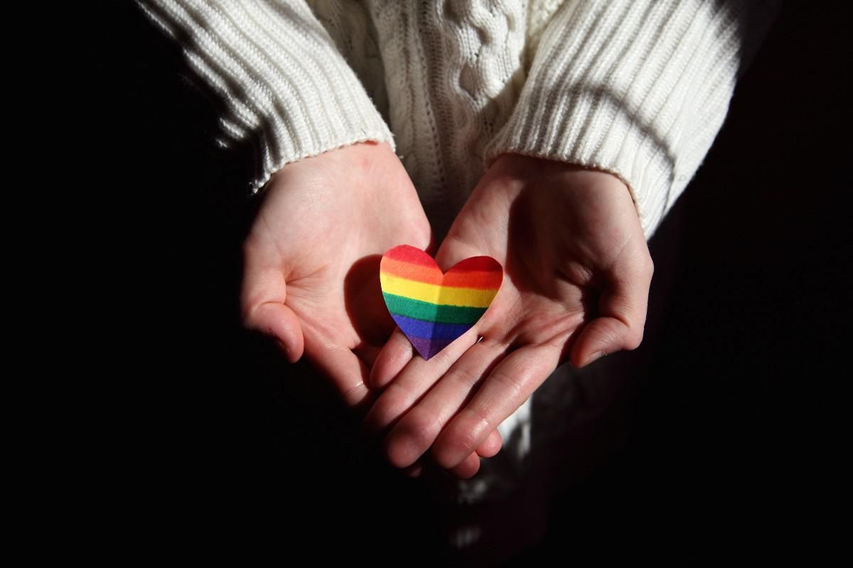 Paper heart with LGBTQ+ flag on it is held in a person's open hands.