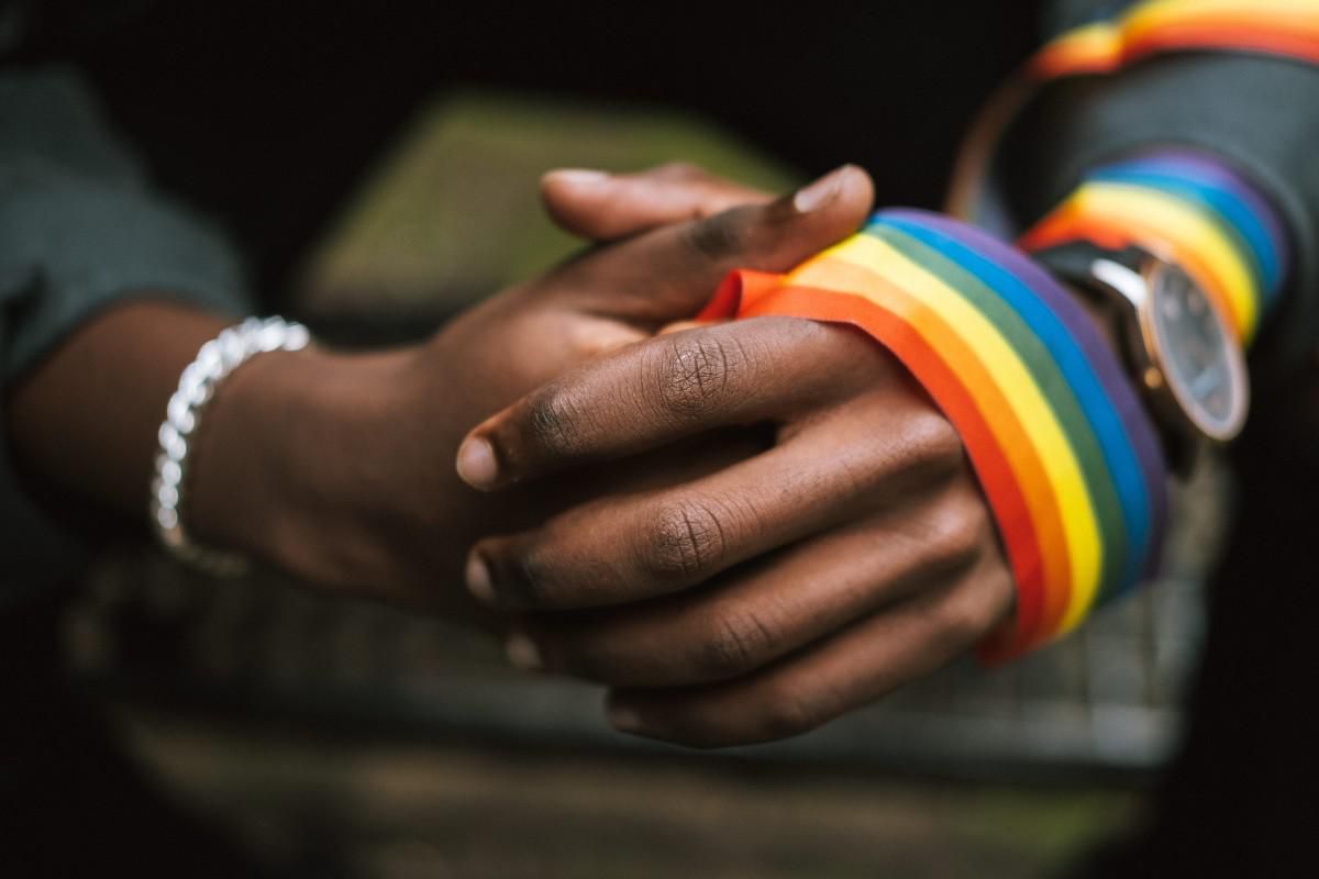 Two POC hands clasped together with a small pride flag wrapped around one of them.