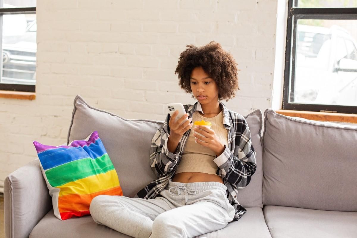A POC sits on a grey couch. They are holding a cup of coffee and looking at their smartphone. A cushion with the pride flag on it is next to them.
