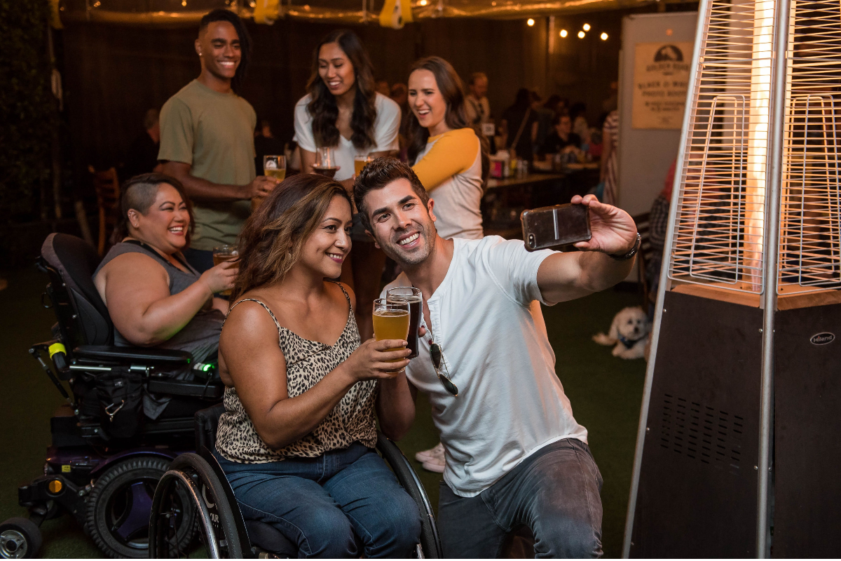 Group of people at a bar having drinks and taking pictures. Two of the people are in wheelchairs.