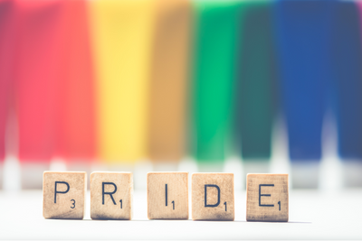The word pride on scrabble letter squares against a faded rainbow background. 
